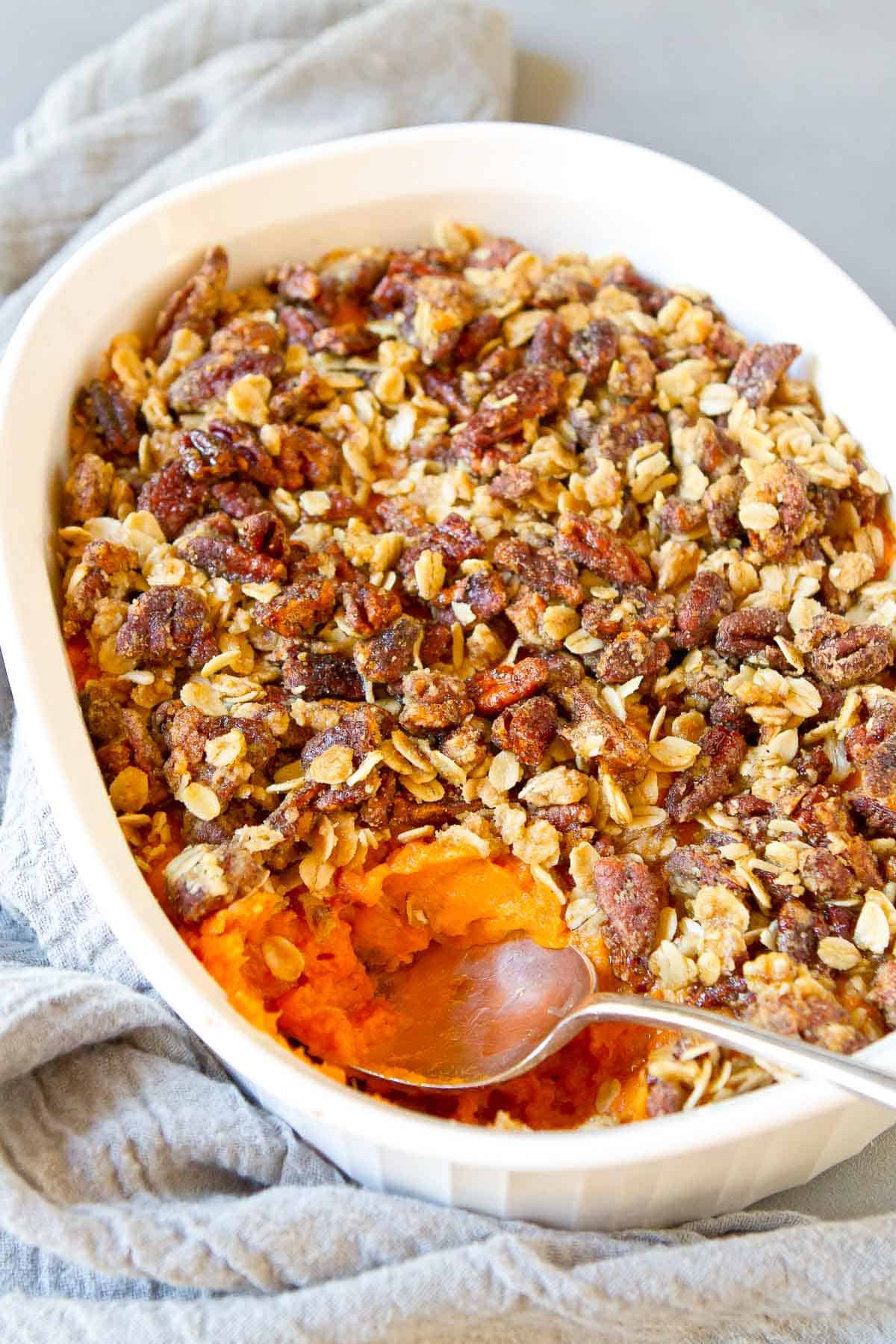 Sweet potato casserole with pecan topping in a white casserole dish, with a silver spoon. Gray napkin underneath.