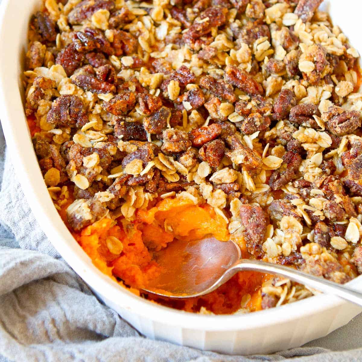 Sweet potato casserole with pecan topping in a white casserole dish, with a silver spoon. Gray napkin underneath.