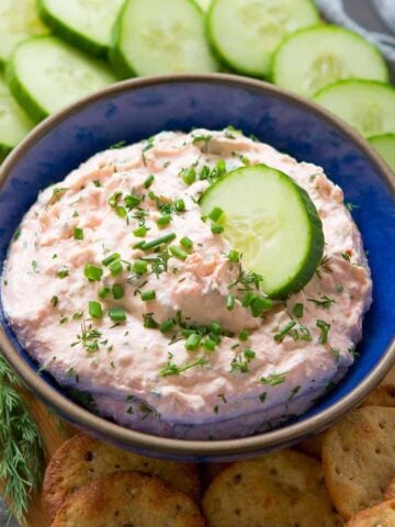 Smoked salmon dip in a blue bowl, surrounded by cucumber slices and crackers.