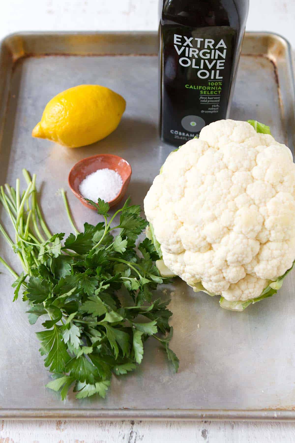 Head of cauliflower, lemon, parsley and olive oil on a baking sheet.