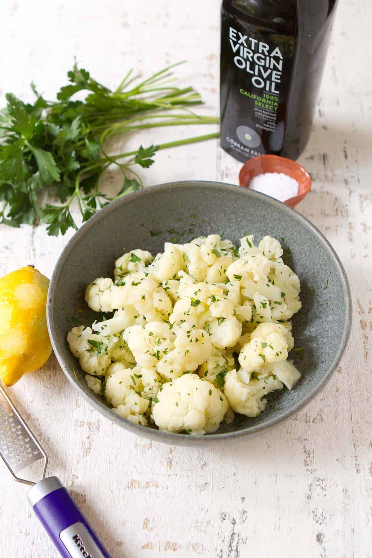 Cooked cauliflower florets in a gray bowl, surrounded by a lemon, parsley and olive oil.