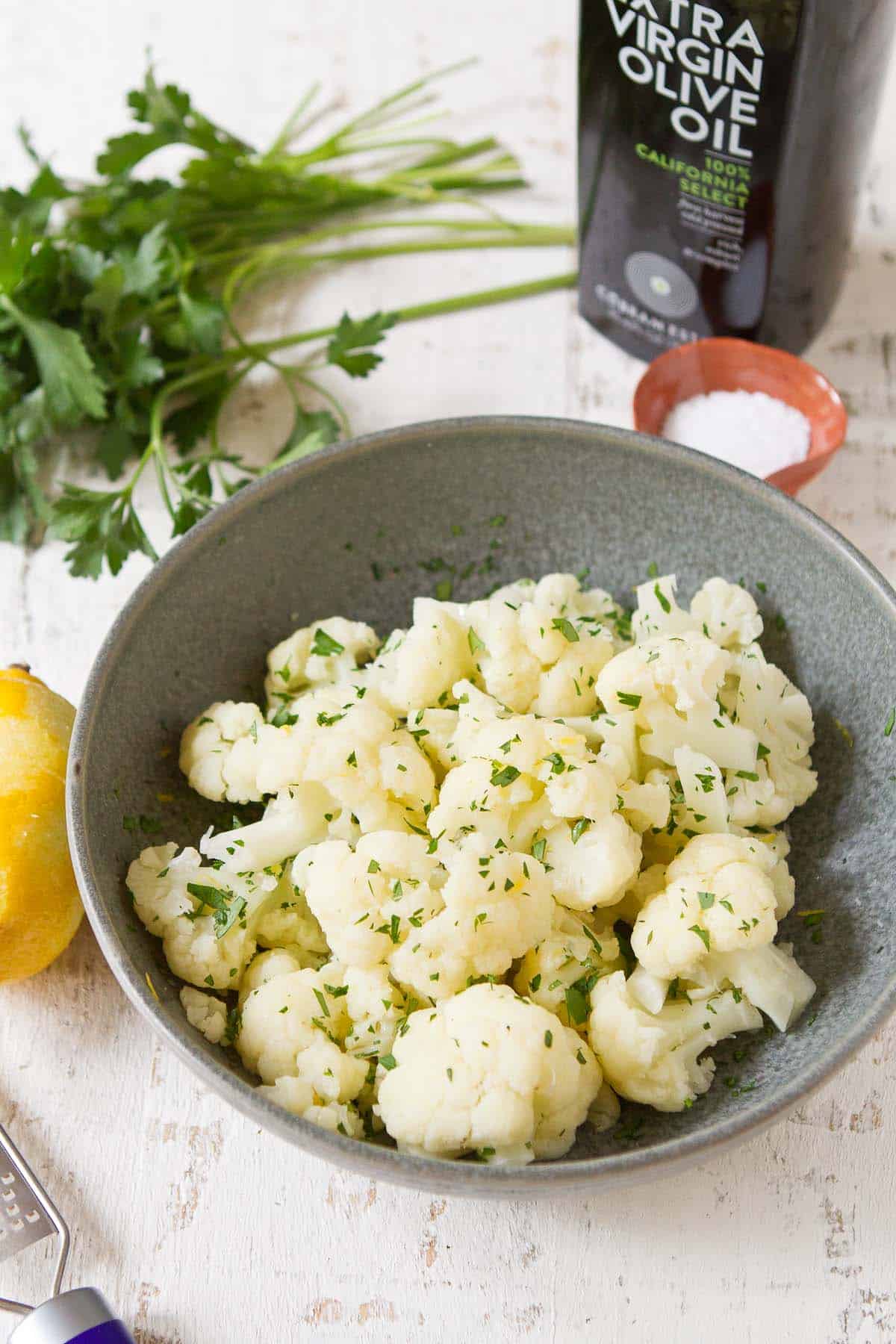 Steamed cauliflower in a gray bowl, with parsley and lemon behind.