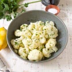 Steamed cauliflower in a gray bowl, with parsley and lemon behind.