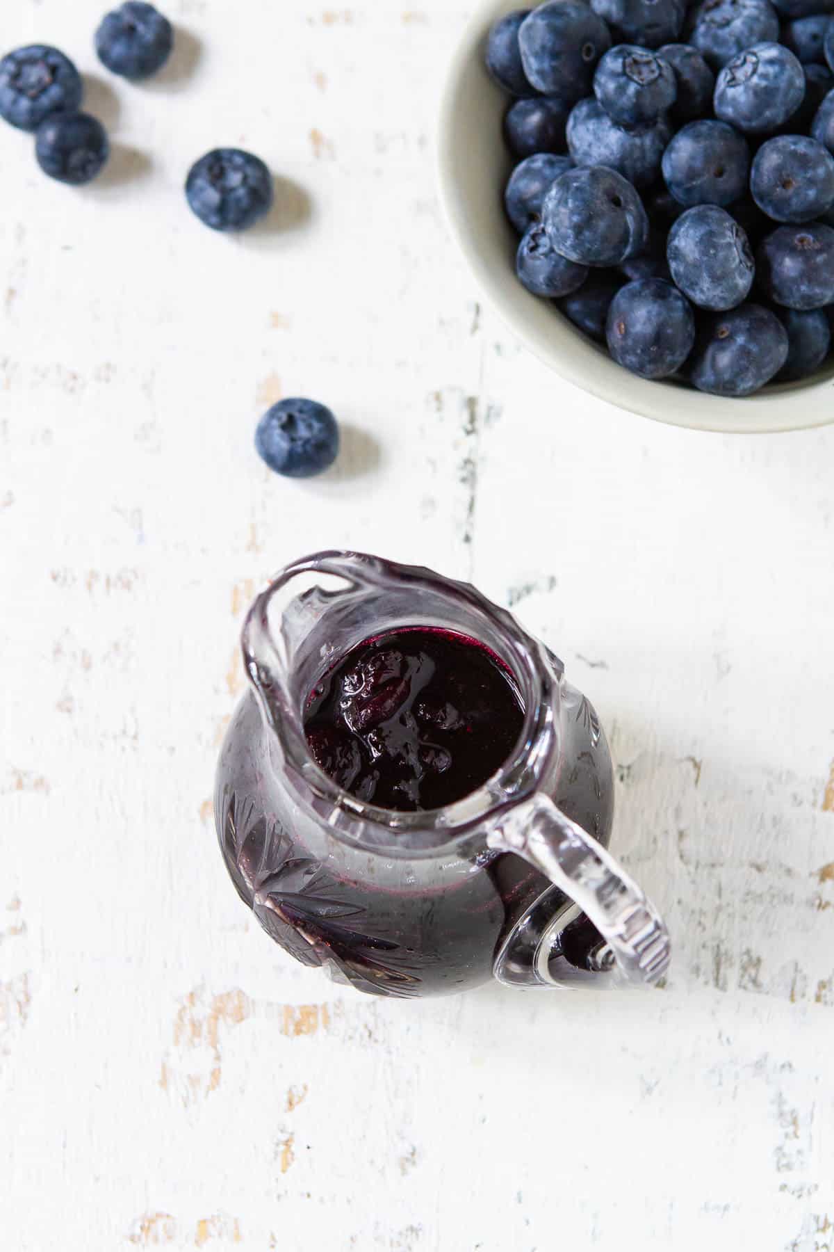 Maple blueberry syrup in a glass pitchers and blueberries in a white bowl.
