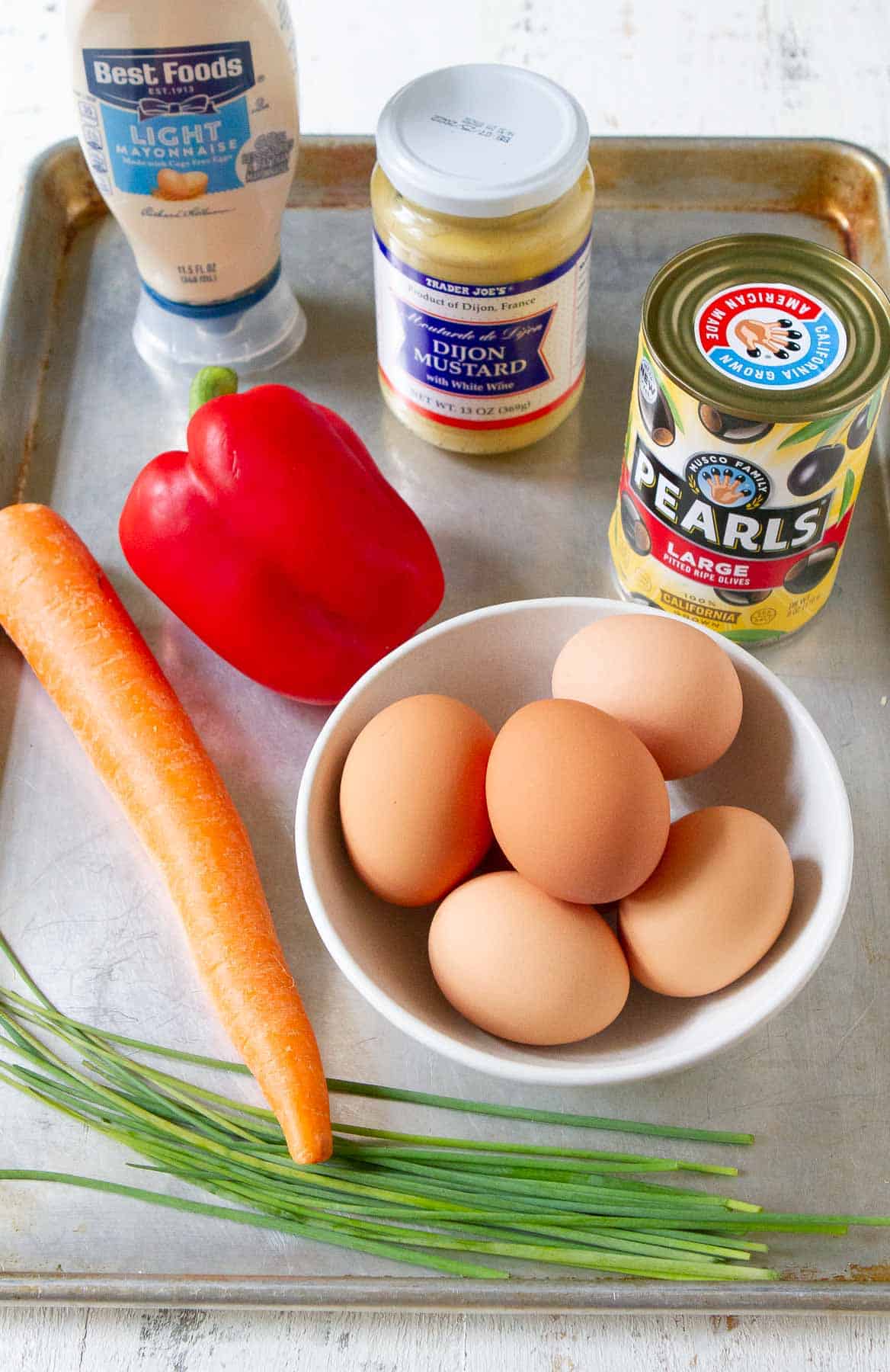 Eggs, mayonnaise, mustard, black olives, carrot, chive and bell pepper on a baking sheet.