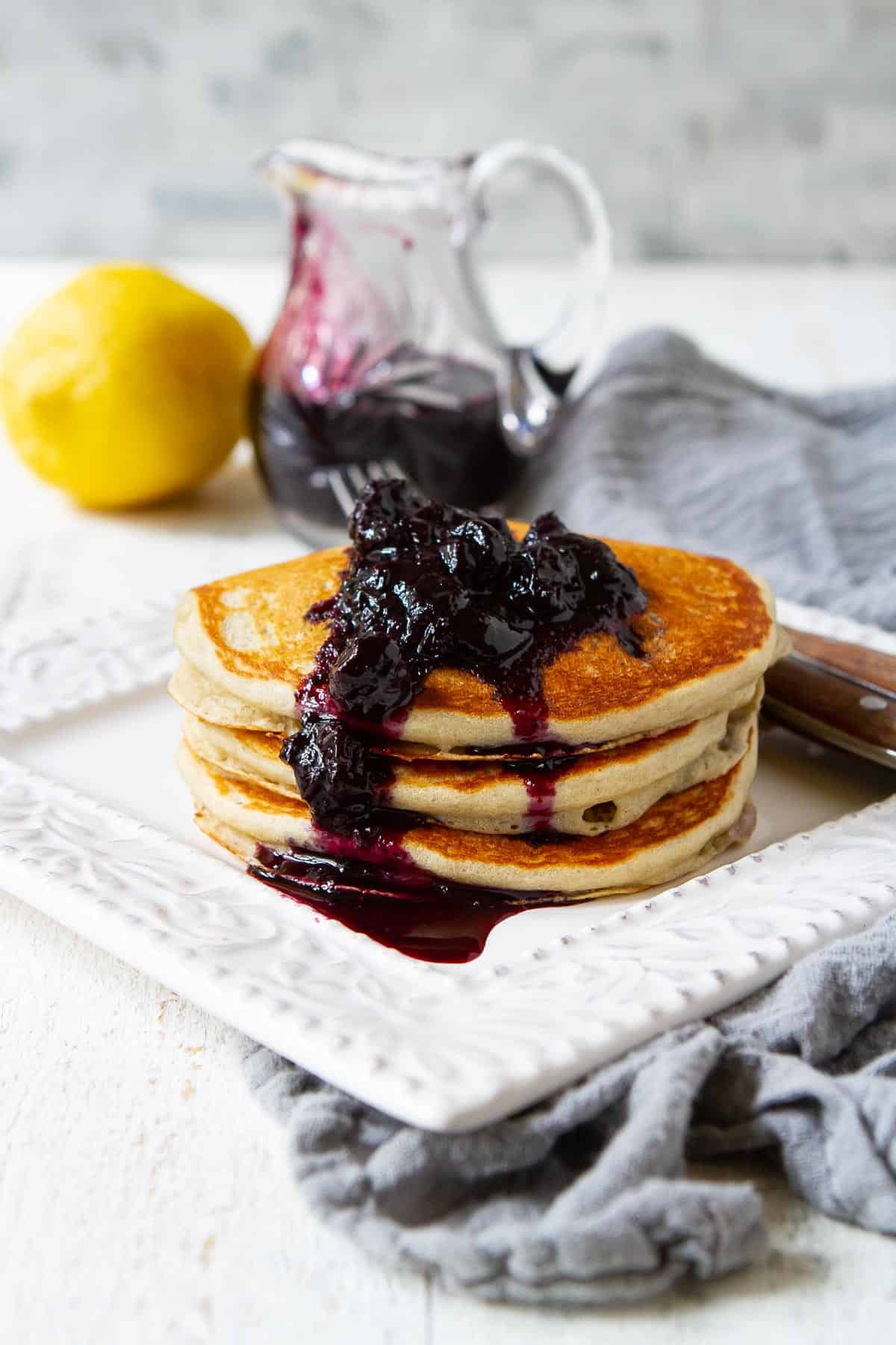 Blueberry lemon pancakes on a white plate, topped with blueberry syrup. Pitcher of blueberry syrup behind.