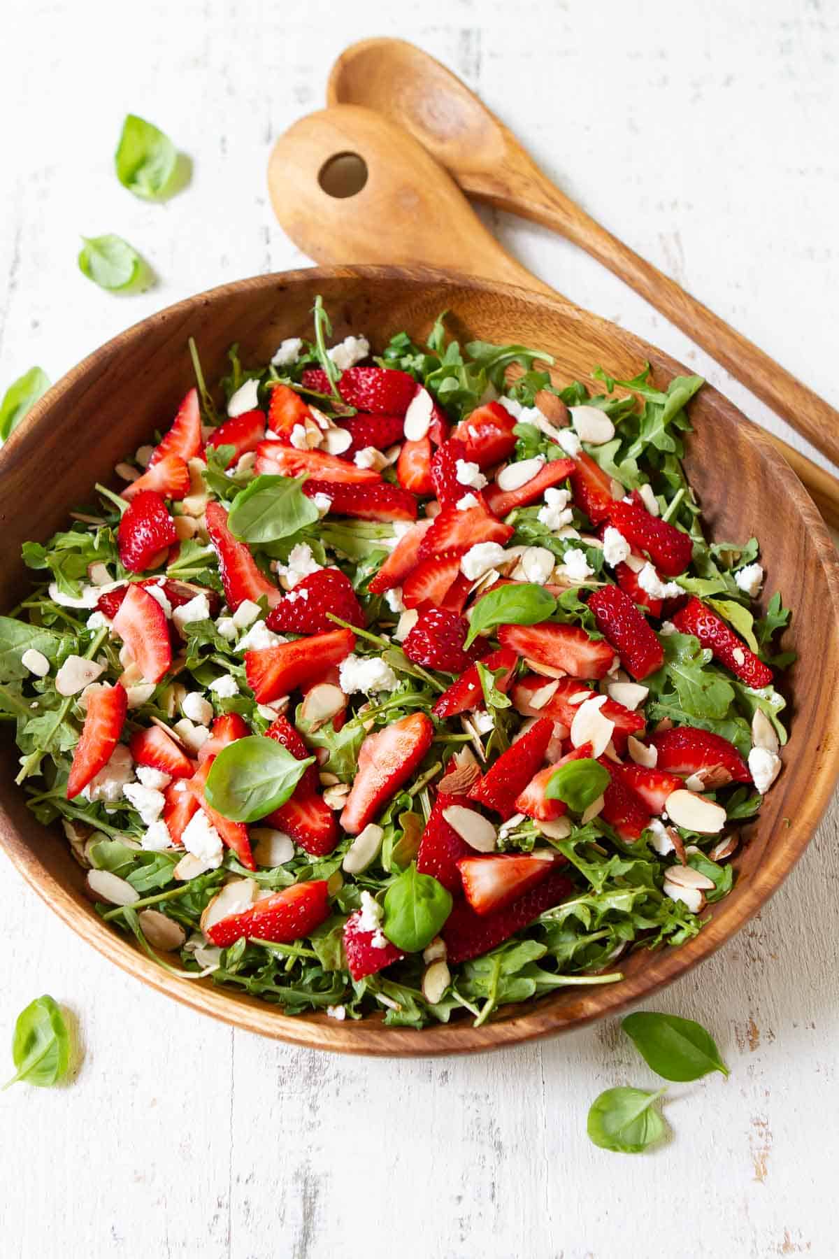 Overhead photograph of arugula salad with strawberries, cheese and almonds in a wooden bowl.