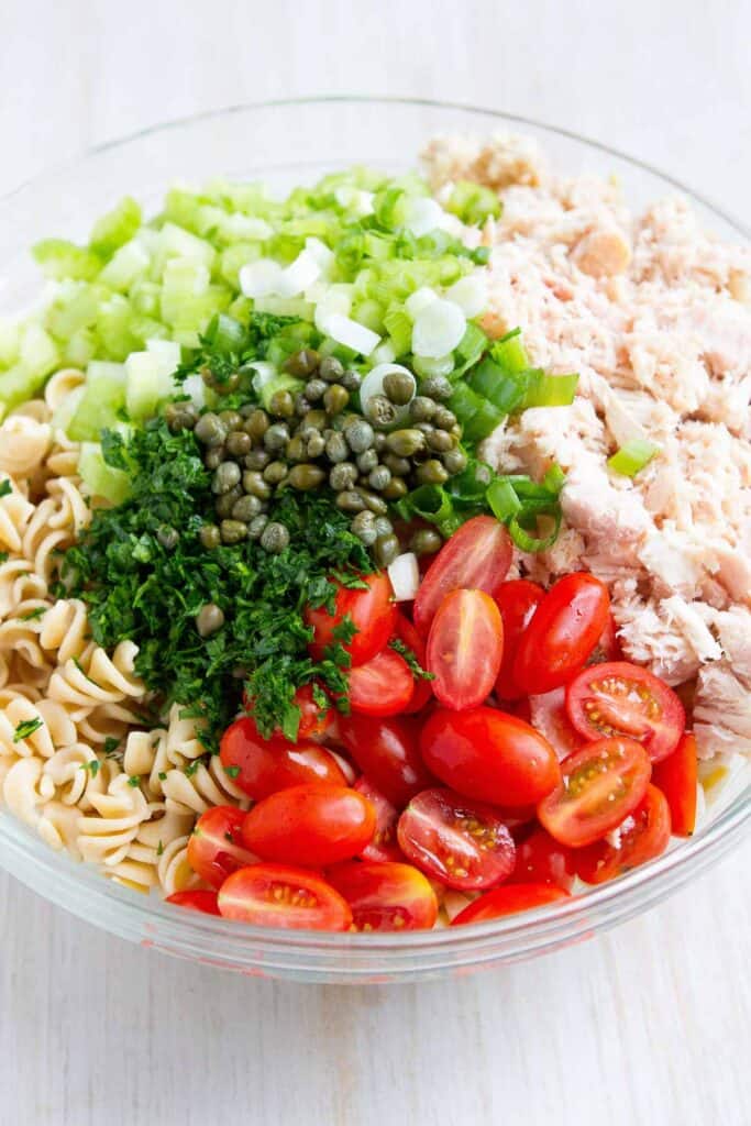 Whole wheat pasta, tuna, tomatoes, celery, capers, parsley and green onions in a large glass bowl.