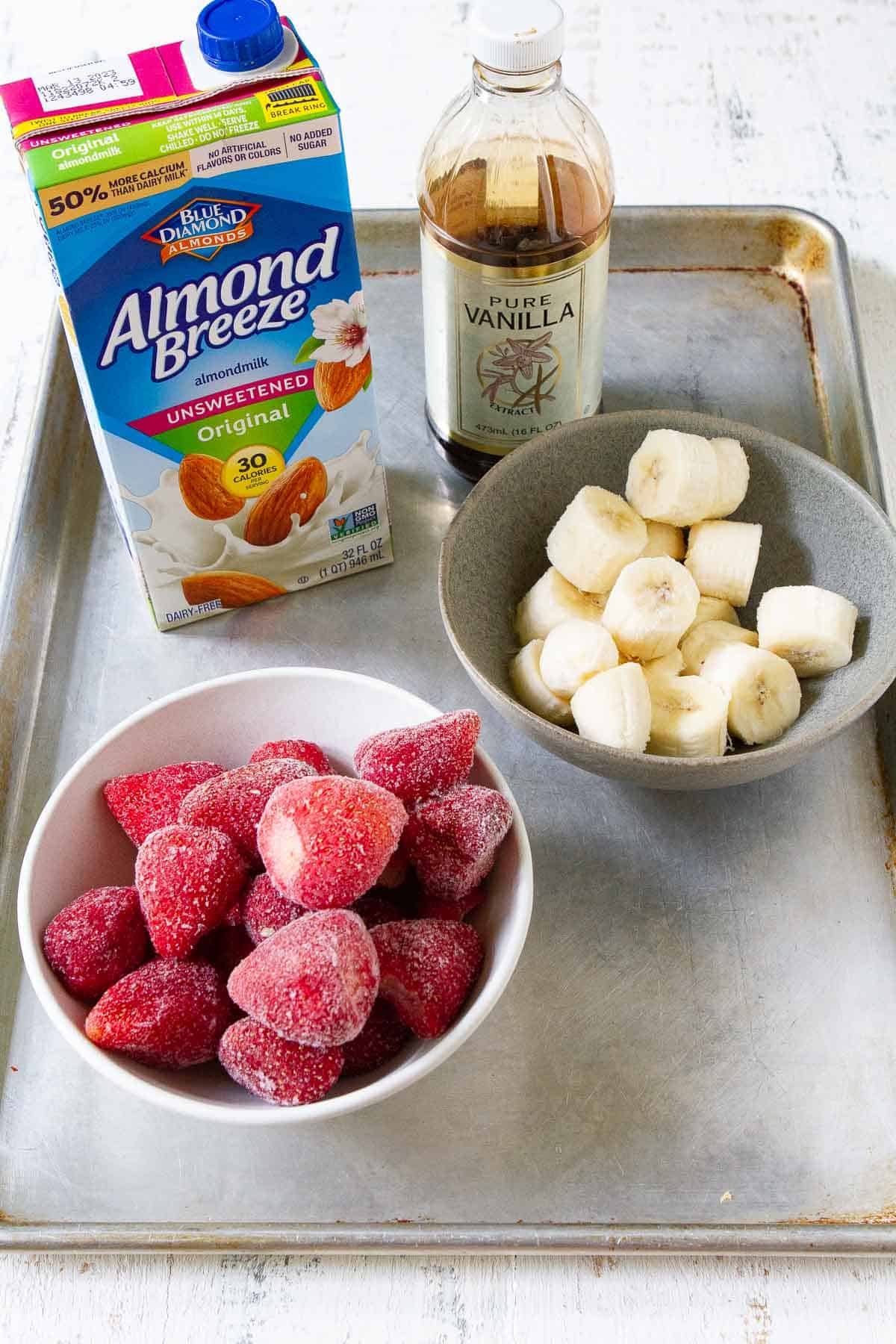 Bowls of strawberries and bananas, vanilla extract and almond milk on a baking sheet.