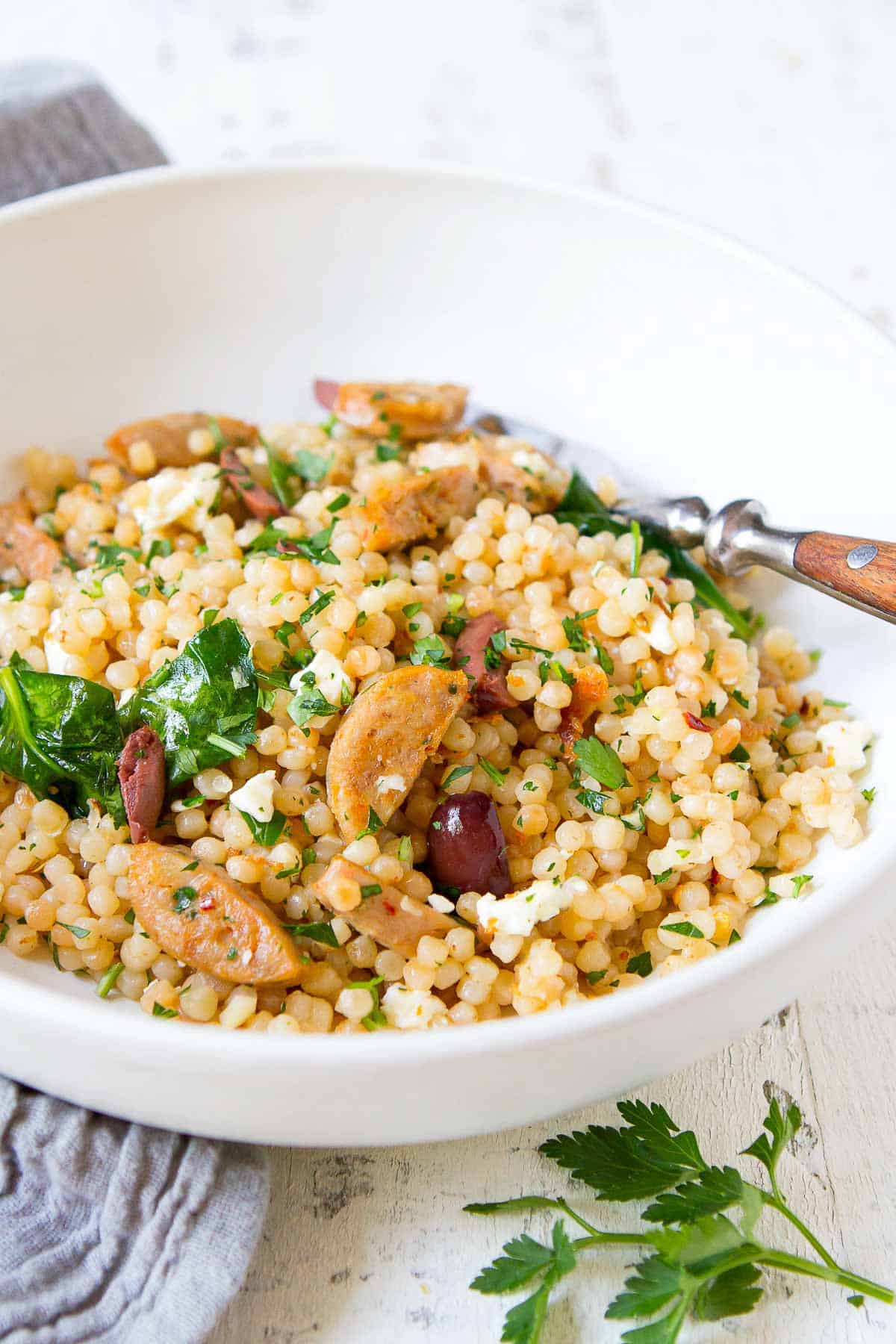 Israeli couscous, cooked sausage, spinach, olives and feta in a white bowl.