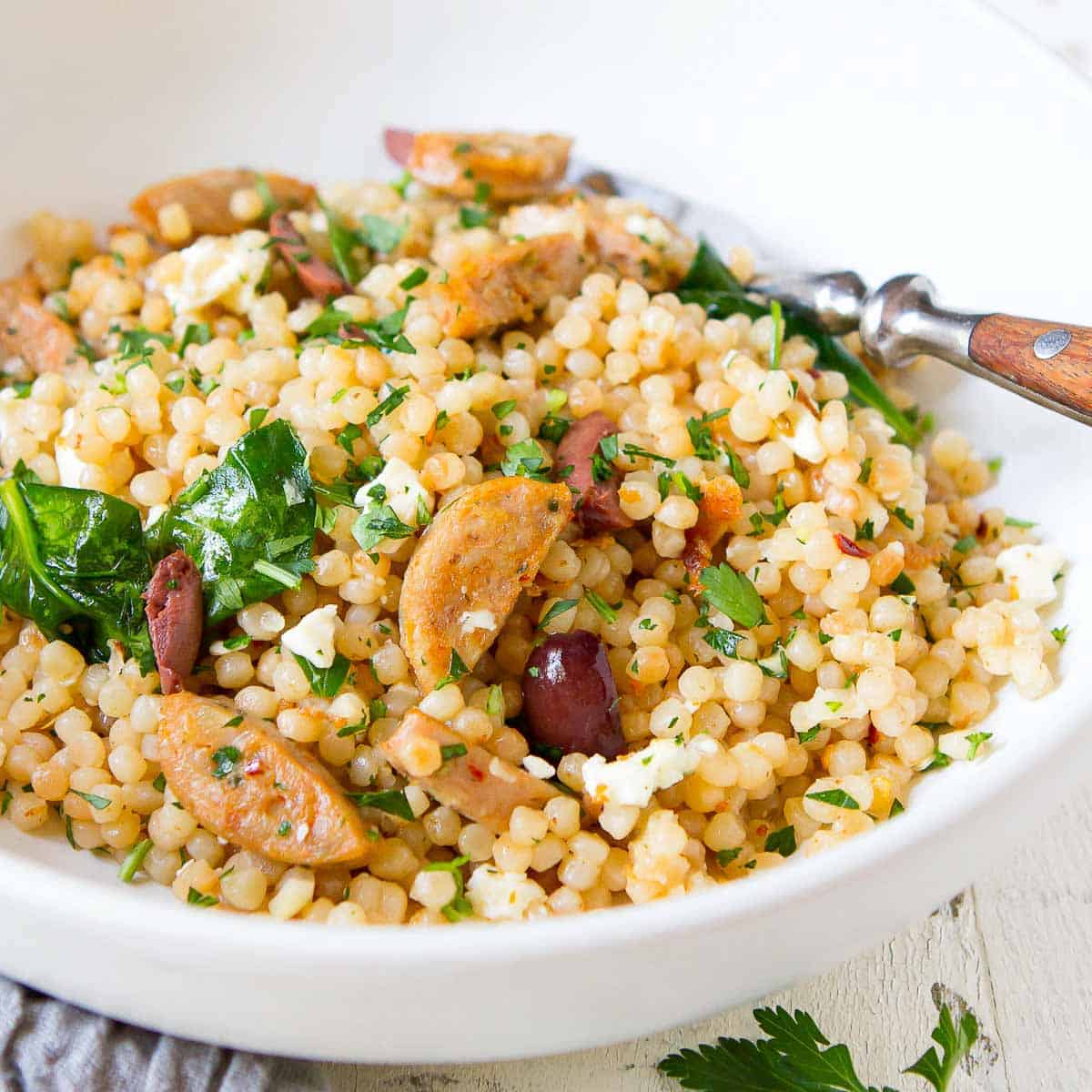 Israeli couscous, cooked sausage, spinach, olives and feta in a white bowl.