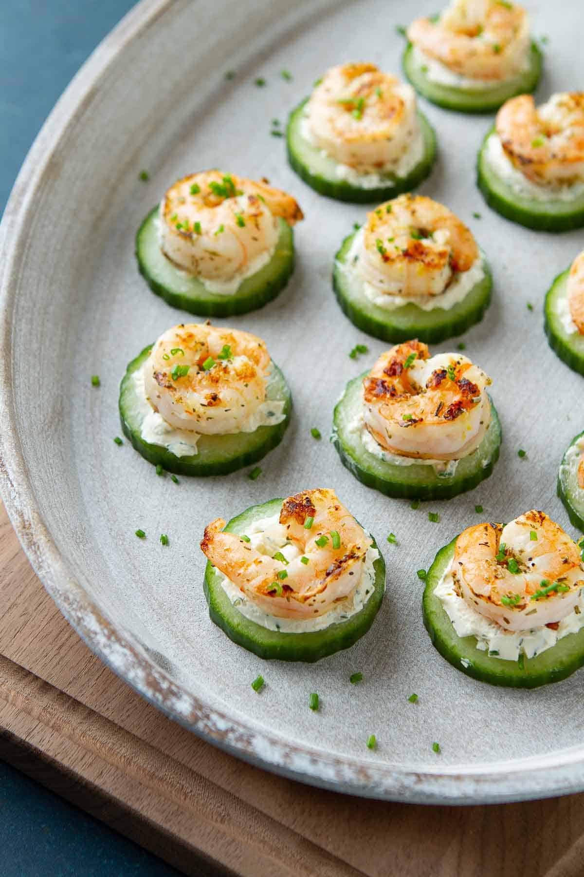 Cucumber slices topped with cream cheese and cooked shrimp, all on a white wooden plate.