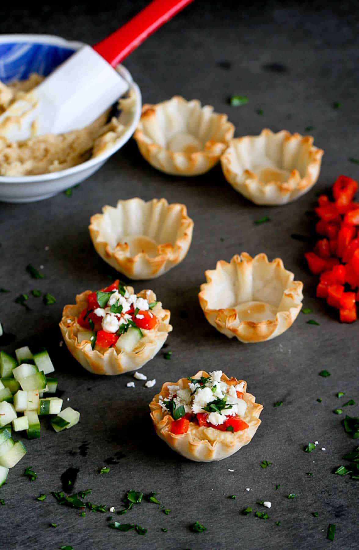 Phyllo shells, partially filled with hummus and vegetables, on a black cutting board.