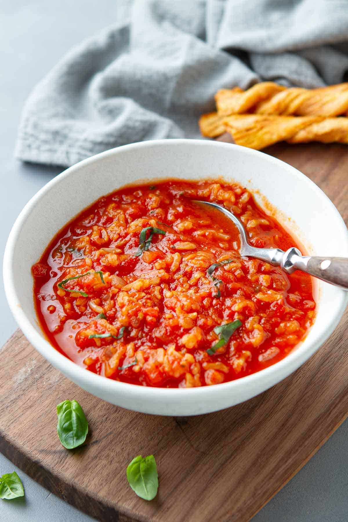 Bowl of tomato rice soup with a spoon, resting on a wooden board, with napkin behind.
