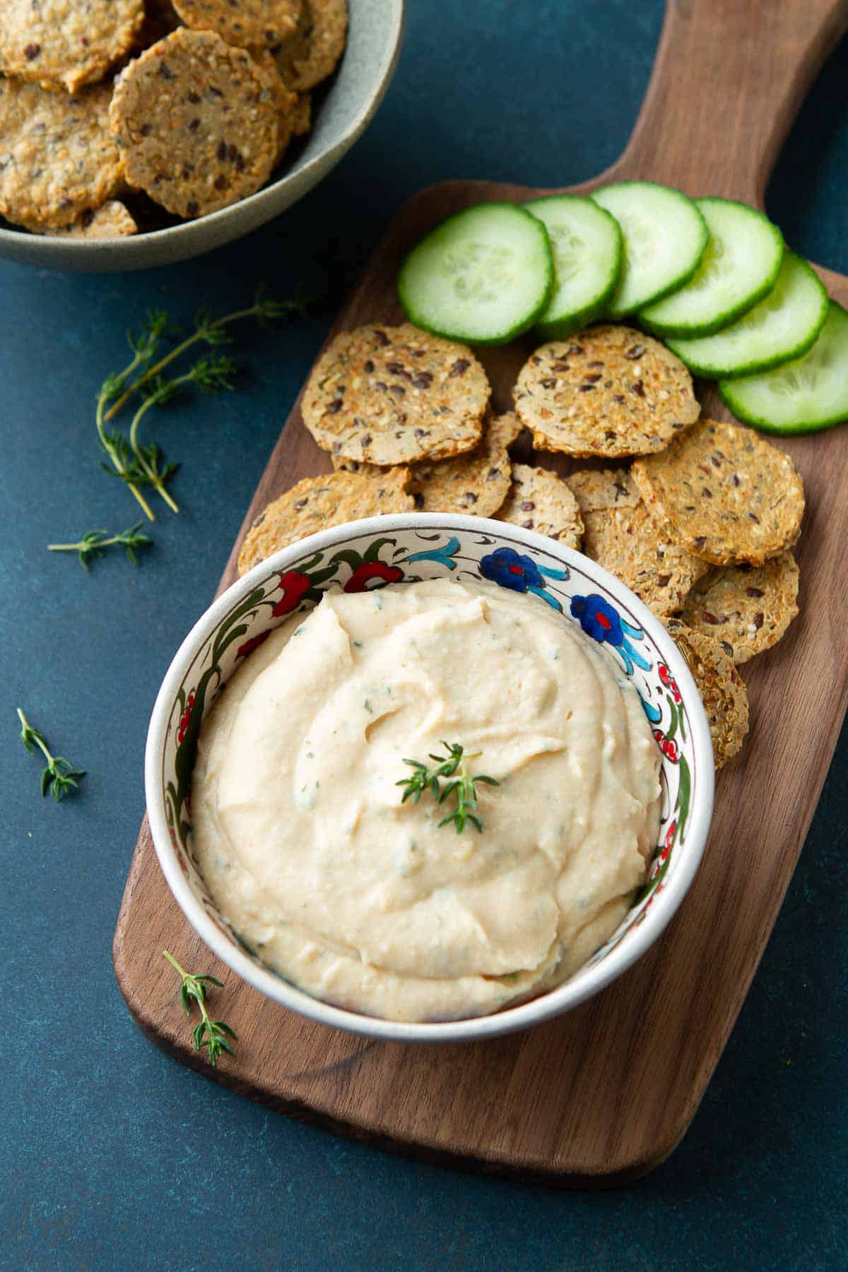 Bowl of white bean hummus, crackers and cucumber slices on a wooden board.