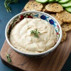 White bean in a bowl, sitting on a wood board with crackers and cucumber slices.