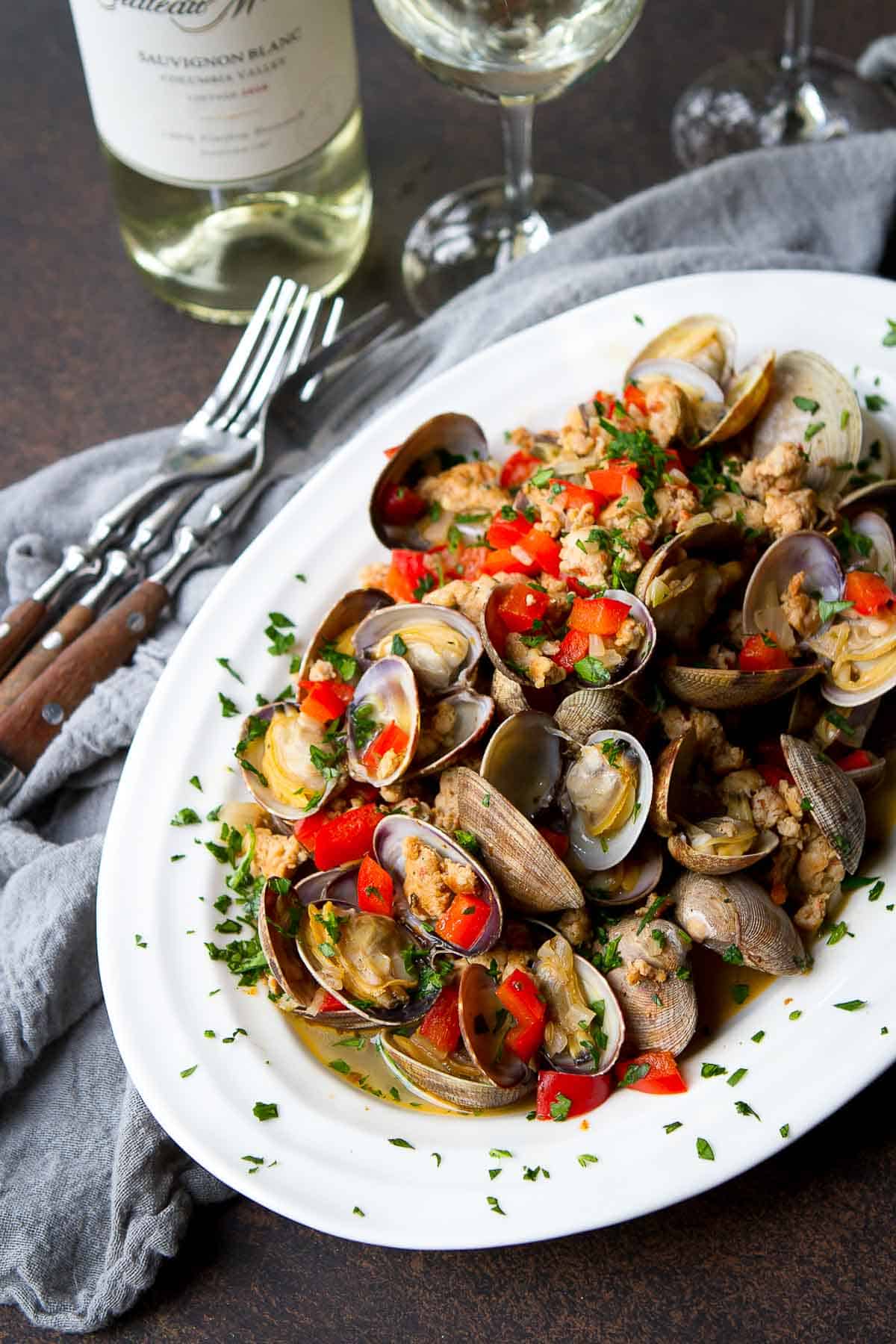 Steamed littleneck clams and sausage on a white plate, with glasses of white wine.