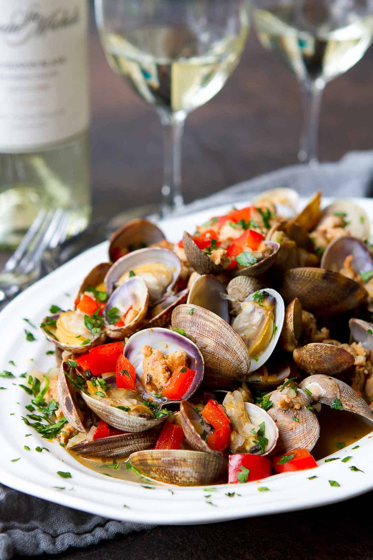 Steamed clams, sausage and red pepper in a white bowl, with glasses of wine in background.