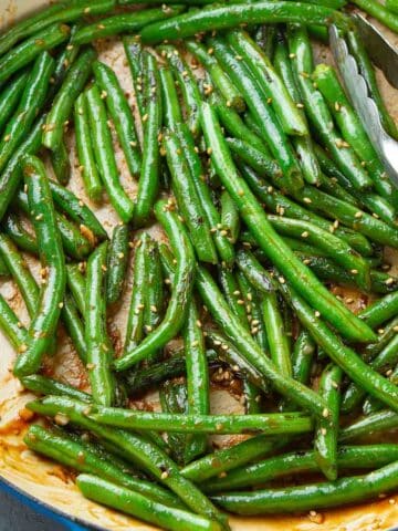 Stir-fried glazed green beans in a large ceramic skillet, with tongs.