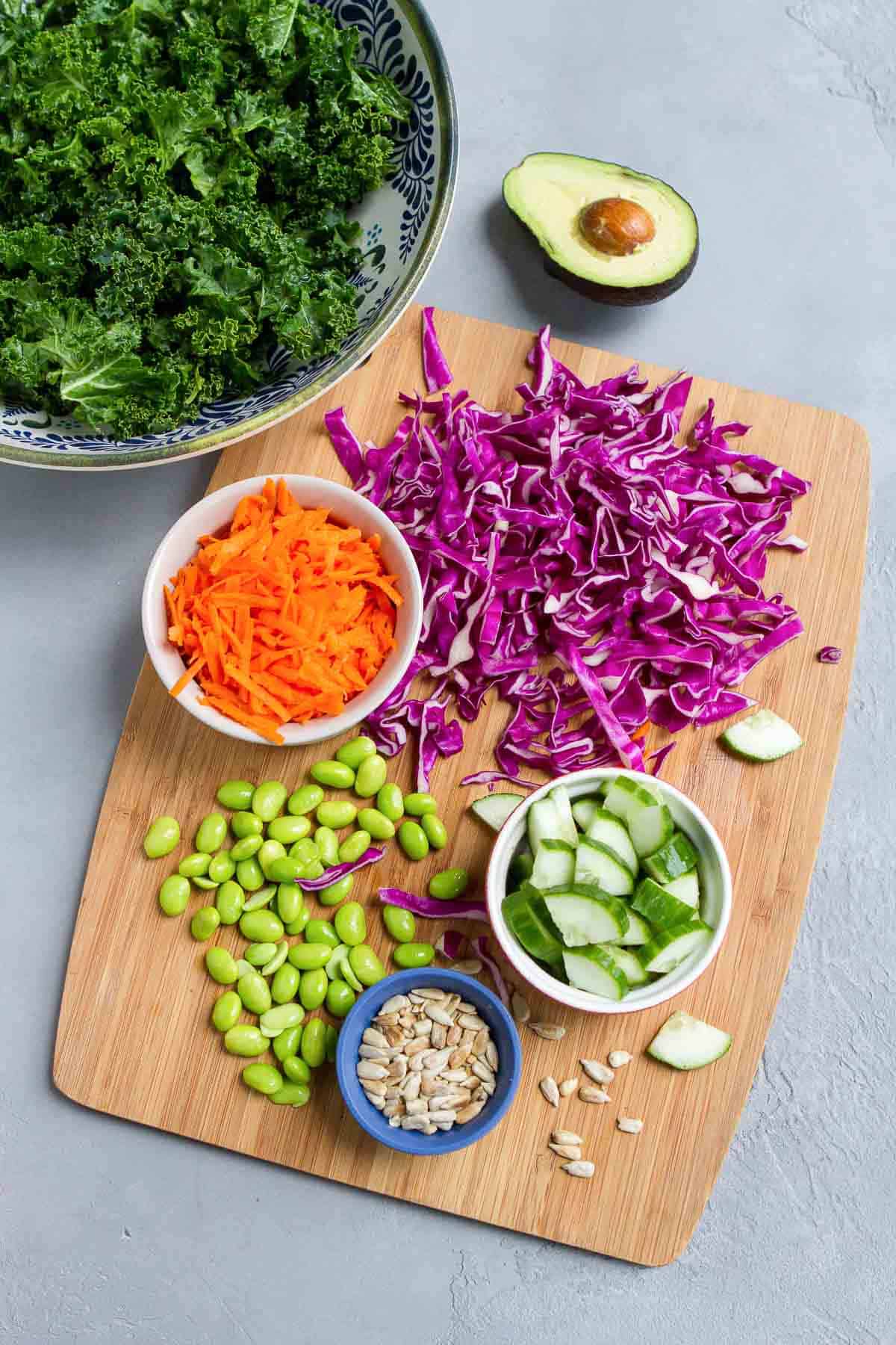 Kale, cabbage, grated carrots, cucumber, edamame, avocado, sunflower seeds and dressing on a bamboo cutting board