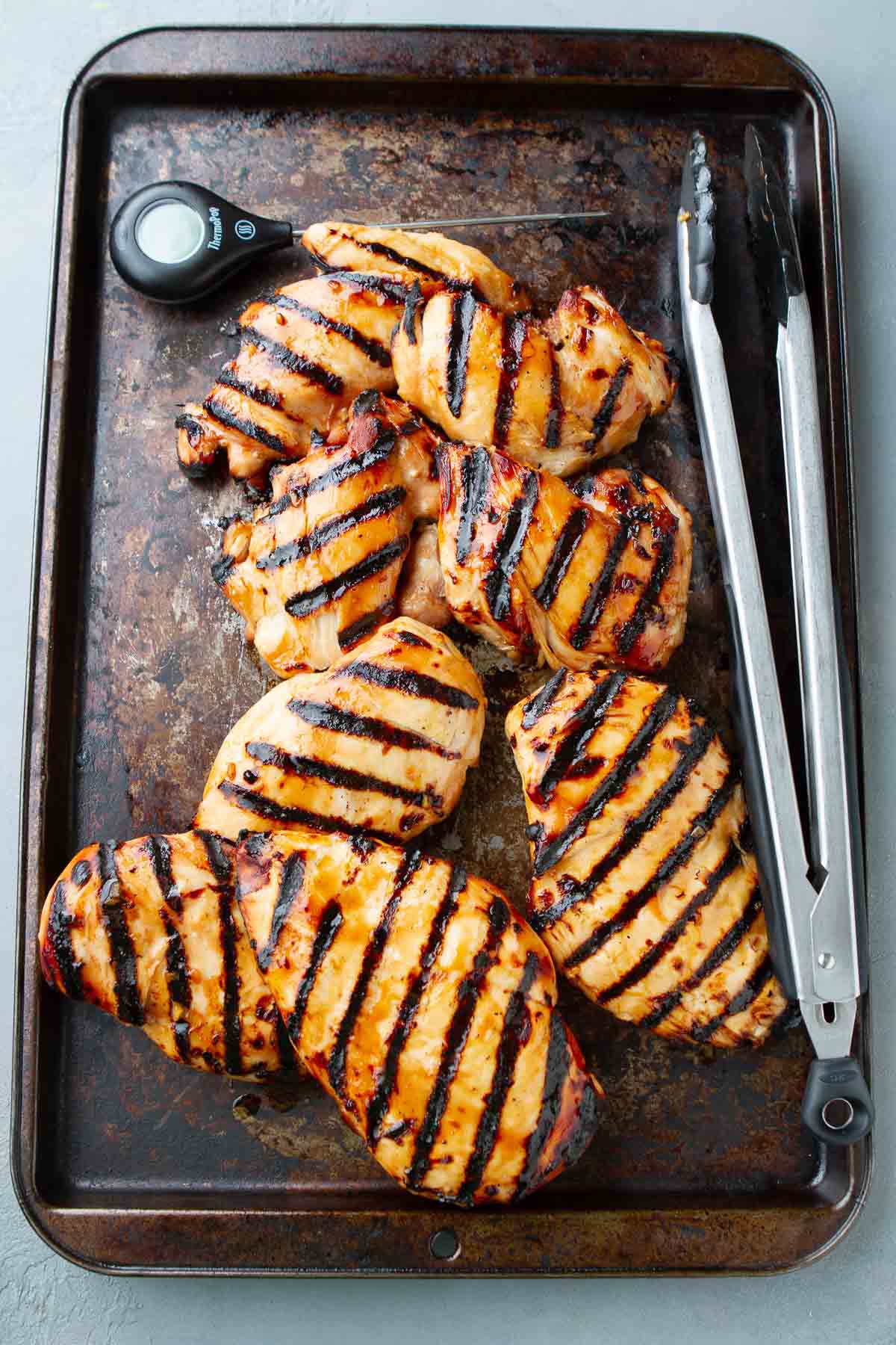 Grilled chicken breasts, tongs and a thermometer on a baking sheet.