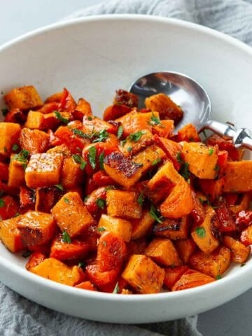 Roasted sweet potatoes and carrots, with a wooden-handled spoon, in a white bowl.