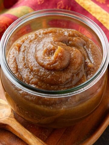 Apple butter in a glass jar, sitting on a wooden tray.