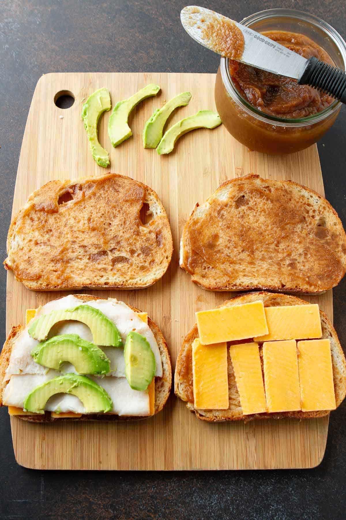 Pieces of bread topped with apple butter, cheese, turkey and avocado, all on a cutting board.