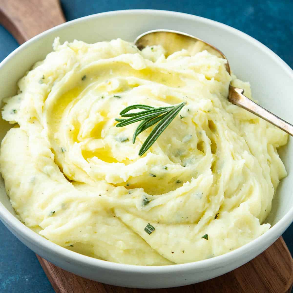 Mashed potatoes with rosemary in a white bowl on a wooden cutting board.