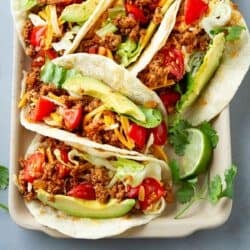 Ground turkey tacos on a platter, topped with avocado slices, tomatoes and grated cheese.