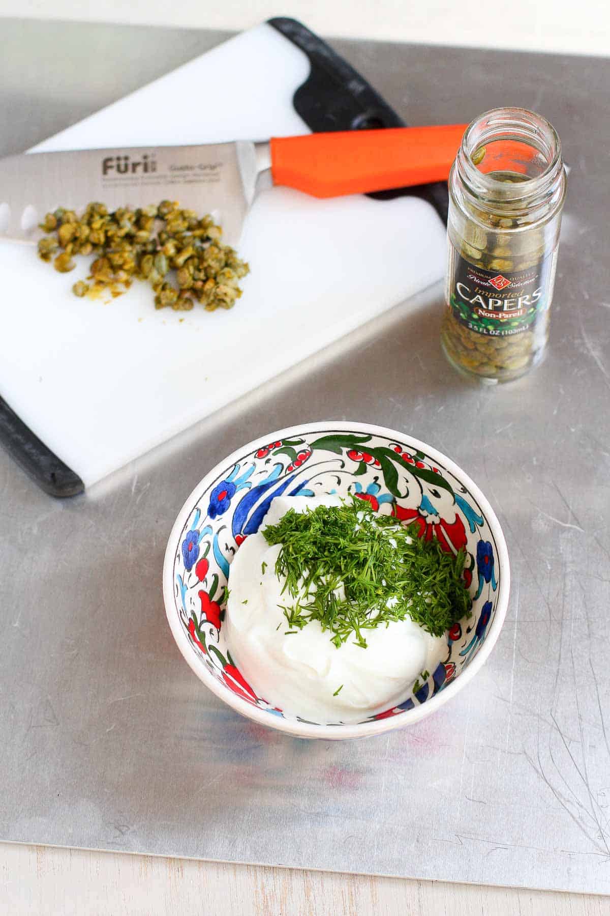 Yogurt and dill in a colorful bowl and chopped capers on a cutting board.
