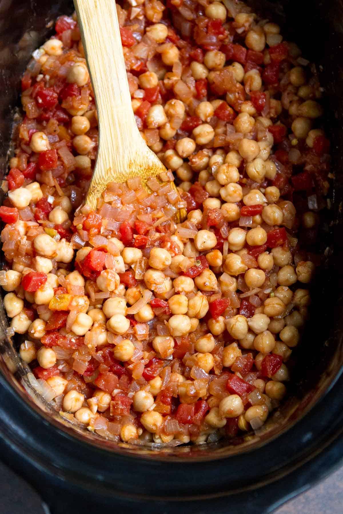 Chickpeas, onions and tomatoes in a crockpot with a wooden spatula.