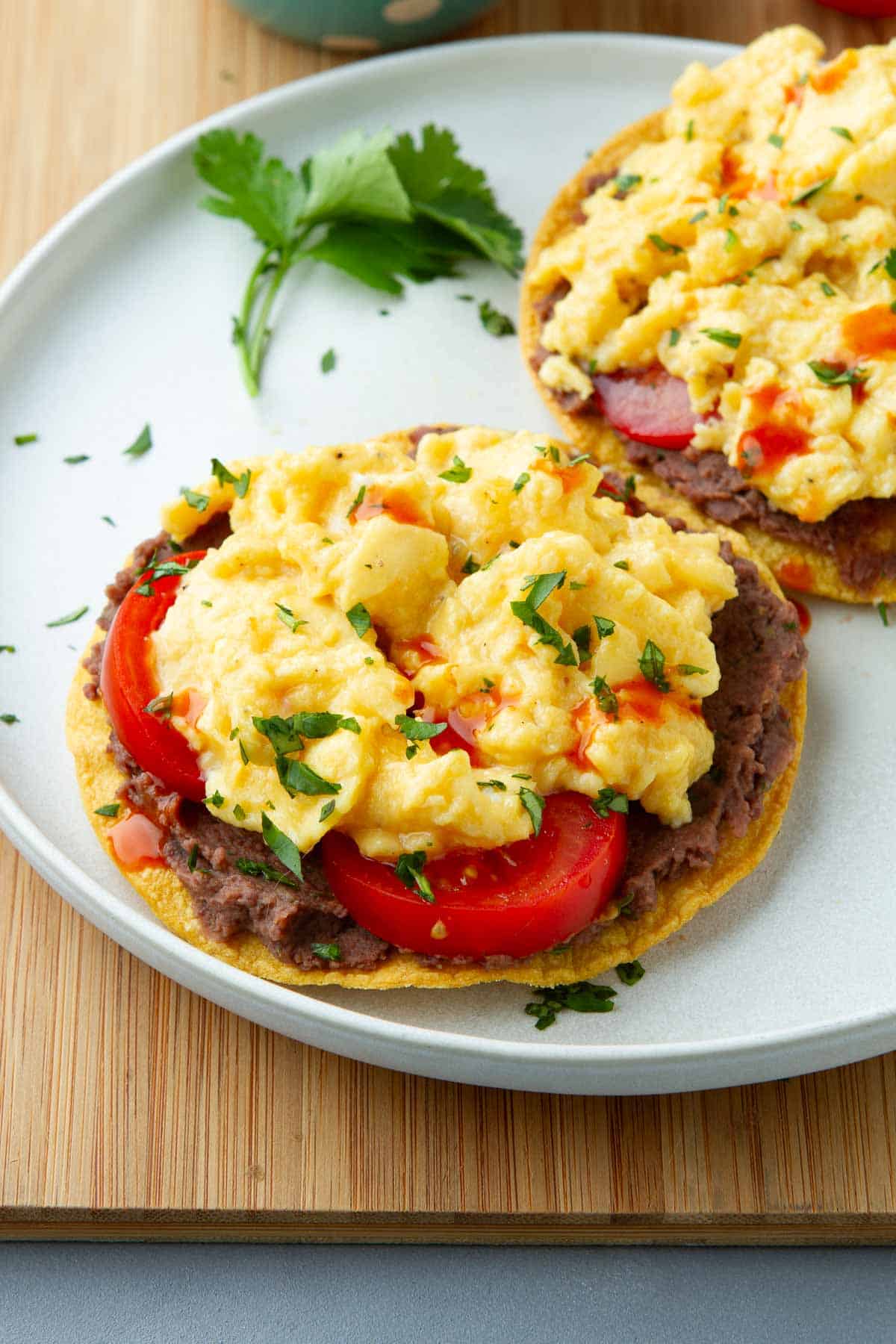Tostadas topped with black bean dip, tomatoes and scrambled eggs on a white plate.