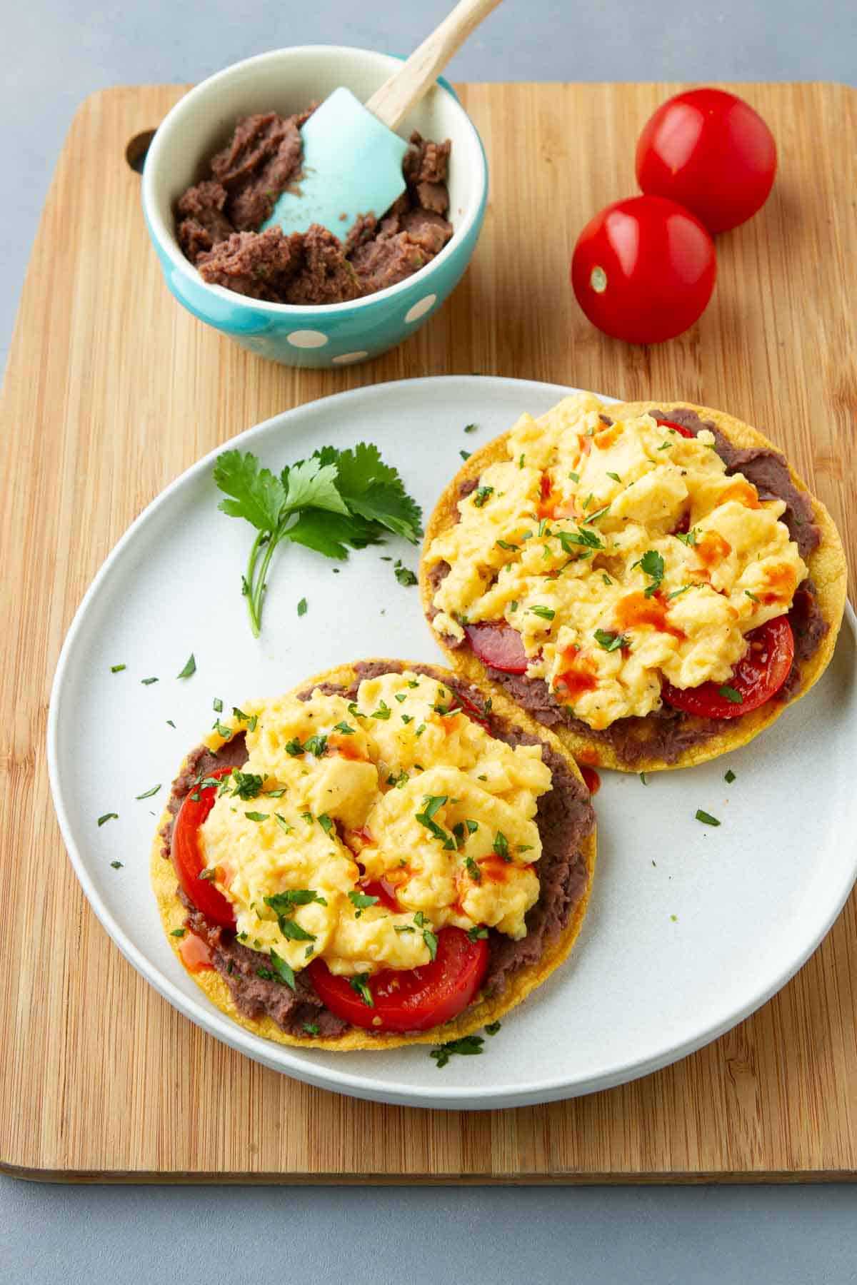 Breakfast tostadas on a white plate, plus a bowl of black bean dip and two tomatoes.